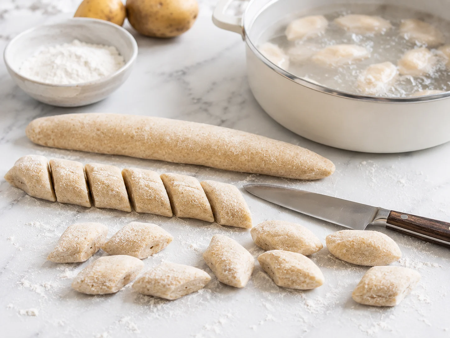 Hands dusted with flour pressing and rolling kopytka dough on a lightly floured work surface — the dough is pale beige with flecks of brown from whole wheat flour, shaped into thick ropes being cut diagonally into small blunt-ended oval pieces roughly three centimeters long, several cut pieces resting nearby showing their characteristic hoof-like flat oval form, a pot of gently boiling salted water visible in the background with a few dumplings floating to the surface