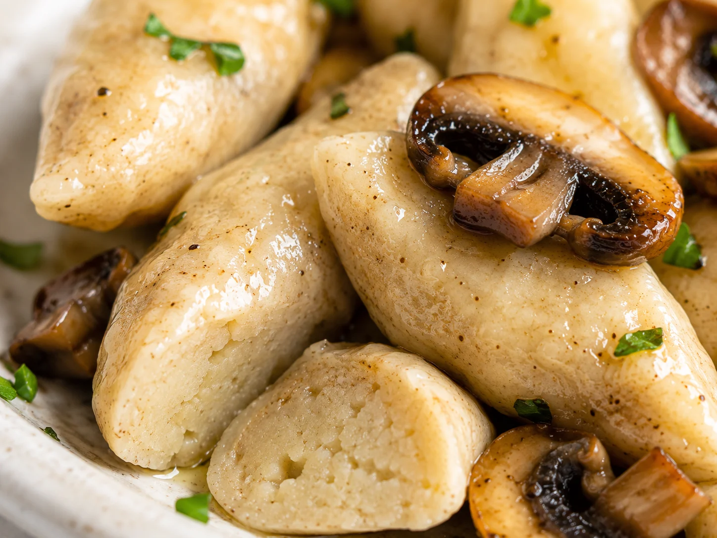 An extreme close-up of three or four kopytka dumplings revealing their soft slightly chewy surface texture with faint whole wheat flour speckling across the ivory-beige skin, their blunt-ended oval hoof shape unmistakable, one dumpling sliced open showing the dense fluffy potato interior, a golden sautéed mushroom slice resting directly on top with olive oil sheen, these are boiled potato dumplings not pierogi not gnocchi not pancakes