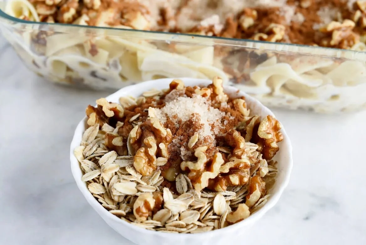 Close-up of a white bowl holding the actual kugel topping mixture of old-fashioned rolled oats, chopped walnuts, sugar, and cinnamon, with a rectangular glass baking dish of wide whole wheat noodle kugel visible in the background before baking.