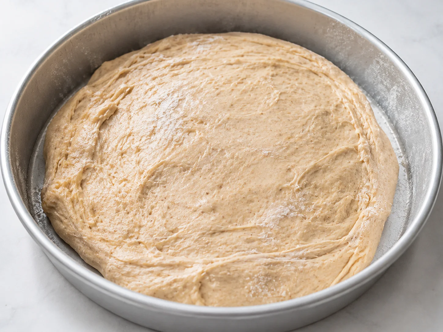 Close-up still-life of a smooth, slightly tacky whole-wheat yeast dough being pressed and spread into a lightly floured round baking pan, its pale golden-beige surface showing the soft elasticity and faint flour dusting of a just-kneaded dough, with no hands present — only the dough itself resting in the pan mid-spread, edges gently pulling inward.