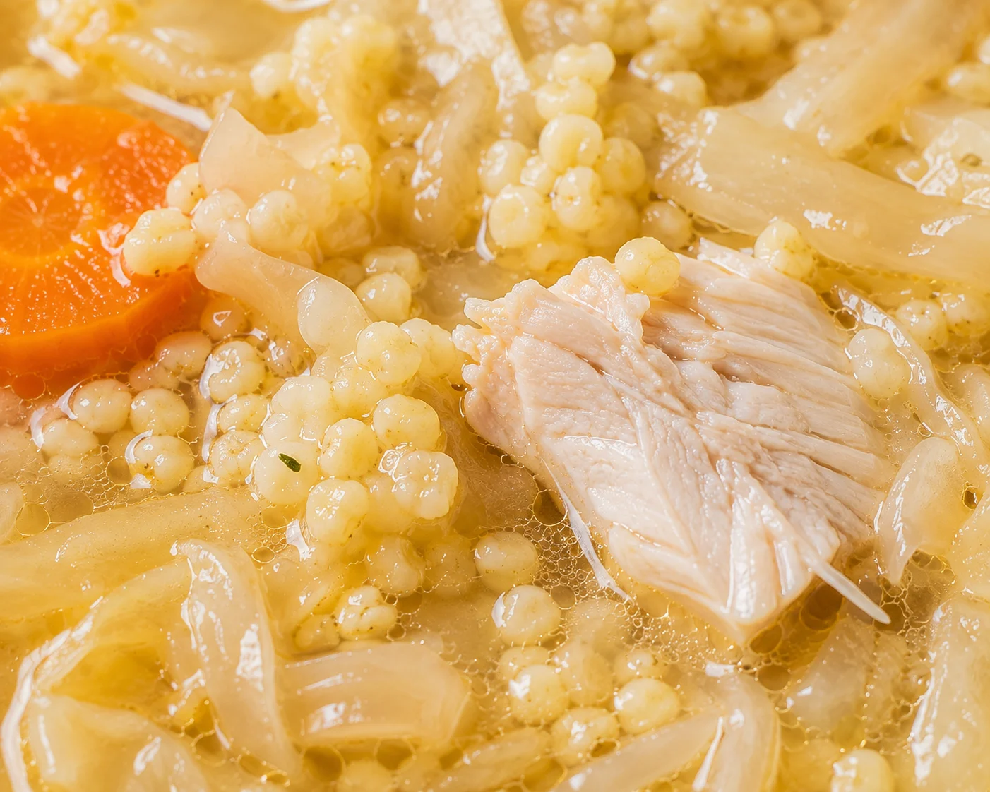 An extreme close-up of the surface of healthy kapusniak in the bowl, revealing the fine texture of swollen millet grains clustered between translucent sauerkraut strands, a glistening golden broth pooling around pale shredded cabbage fibers, a single torn piece of cooked white turkey breast showing its fibrous texture, and tiny droplets of oil catching light on the soup surface