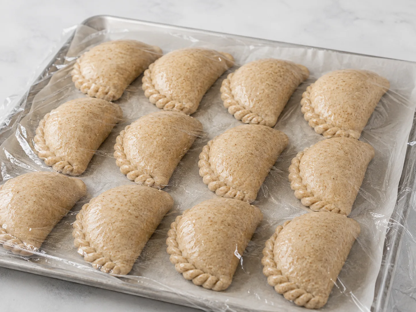 Twelve shaped but unbaked kibinai — half-moon pastries with neatly crimped curved edges made from a whole-grain dough — are arranged on a parchment-lined tray and covered loosely with a piece of lightly oiled plastic wrap, the dough visibly puffy and resting as it proofs, shot in close-up with no people present.