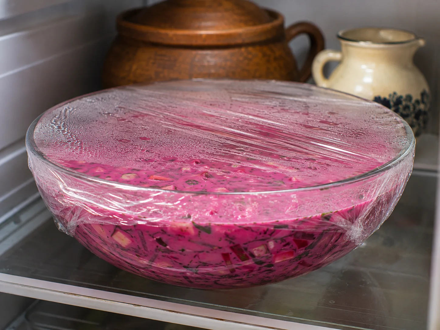 A large bowl of fully combined šaltibarščiai — intensely pink from the grated beets stirred into chilled kefir, with cucumber, scallion, and dill visible throughout — covered tightly with a lid or plastic wrap and photographed in close-up as it rests and chills, condensation just beginning to form on the inside of the cover, the soup's color deep and vivid.