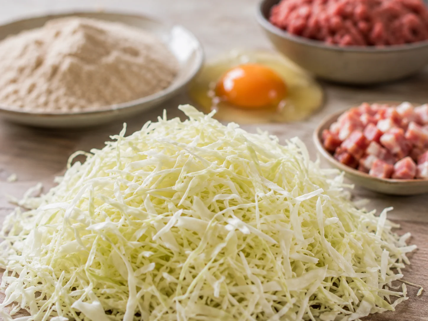A macro close-up on a small mound of finely shredded raw cabbage in sharp focus at the center of the frame, its pale translucent strands just beginning to show the fine texture from fresh shredding, while behind it the rest of the ingredients — a heap of whole wheat flour blended with all-purpose, a cracked egg, a bowl of lean ground beef, and a small dish of diced bacon — recede into soft blurred layers of warm and neutral tones.