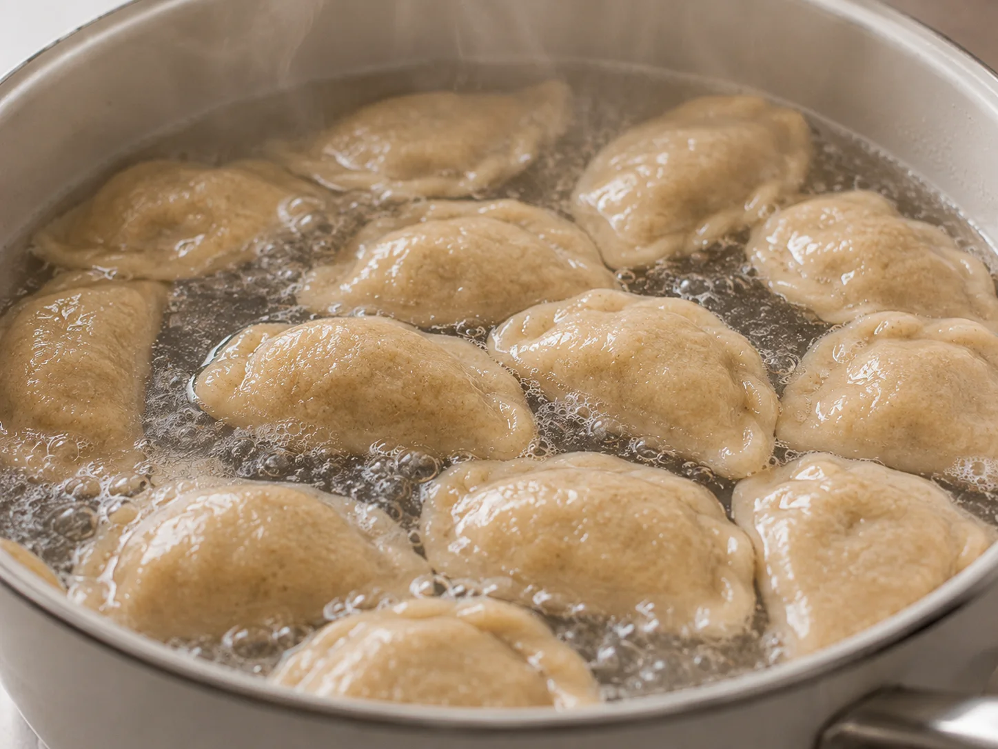 A close-up on a wide pot of generously salted boiling water with a dozen pale half-moon koldūnai dumplings floating near the surface mid-cook, their pinched curved edges plump and tender, gentle wisps of steam curling upward from the water, a few dumplings just beginning to rise while others bob lightly, no people present, the pot rim framing the bubbling water and dumplings in a tight mid-cook moment.
