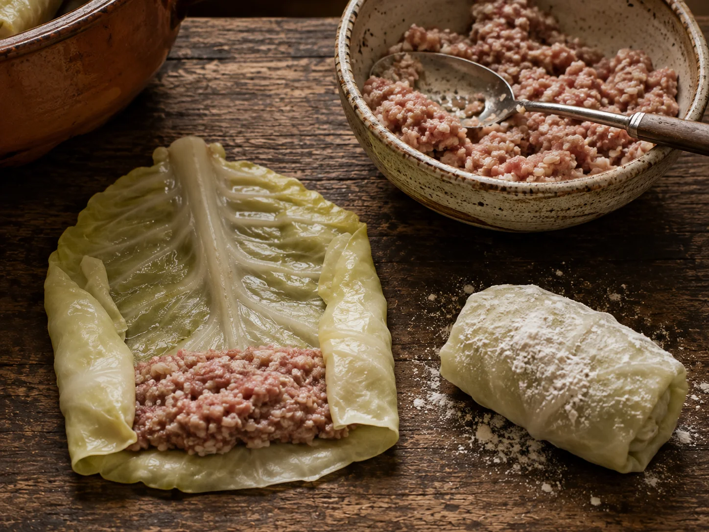 A close-up still life of the rolling step mid-transformation: a softened, pale-green blanched cabbage leaf laid flat with its thick central rib trimmed flush, a generous spoonful of raw ground-pork-and-rice filling pressed onto the lower third, the leaf's two sides already folded inward into a snug parcel, a dusting of all-purpose flour visible on the completed roll resting beside it, and a small bowl of the mixed filling with a spoon resting across it nearby, no person present.