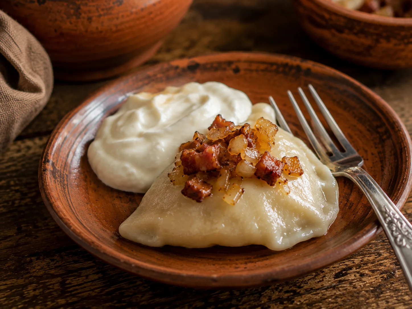 A single small half-moon koldūnai dumpling with a thin, just-boiled egg-dough skin resting on a small plate, topped with a spoonful of crisp diced bacon and softened golden onion with a pool of sour cream beside it, a fork resting against the plate edge in close-up.
